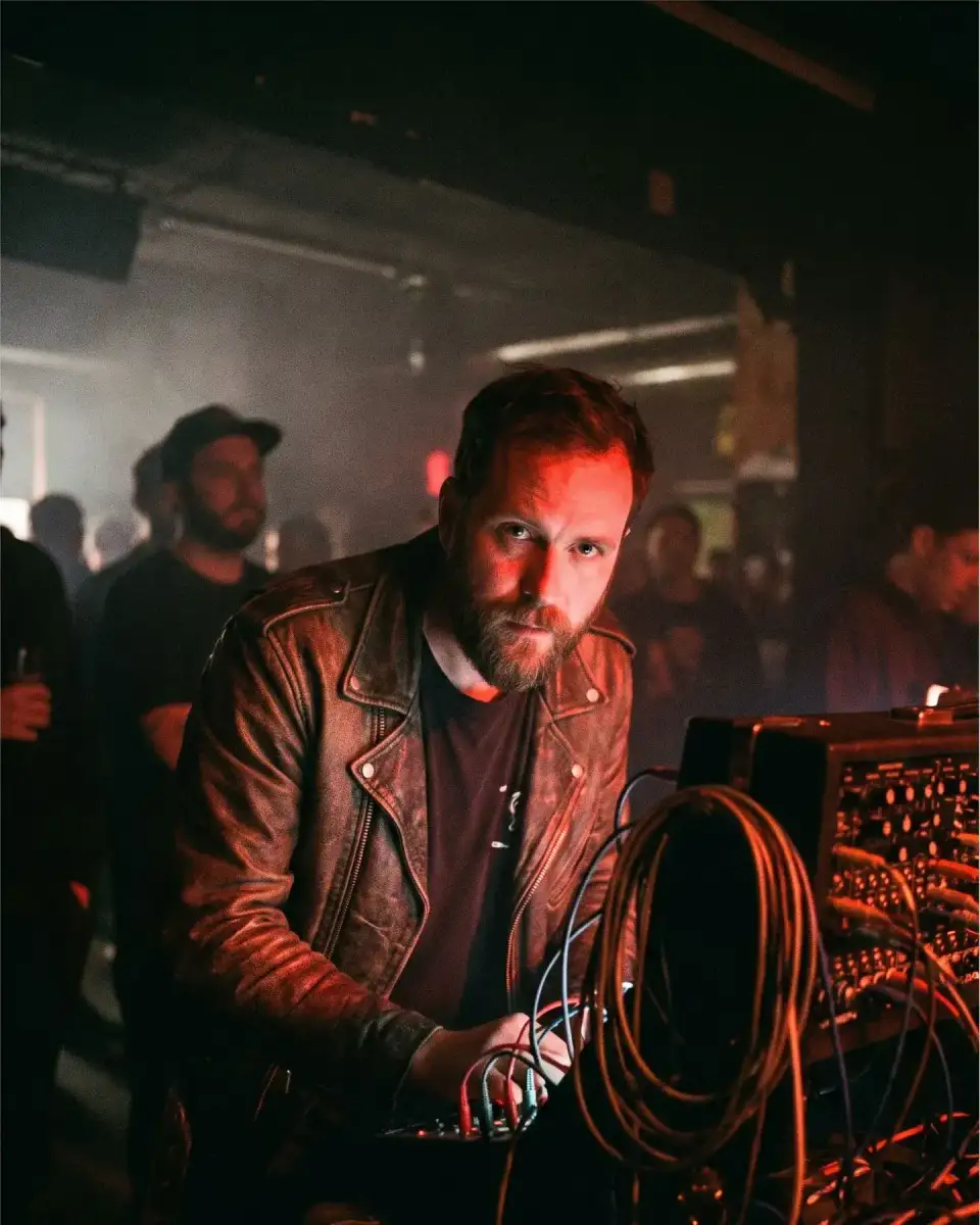 Porträt von Ferhat Demir in Lederjacke hinter einem Modularsynthesizer mit Kabeln, rotes Licht